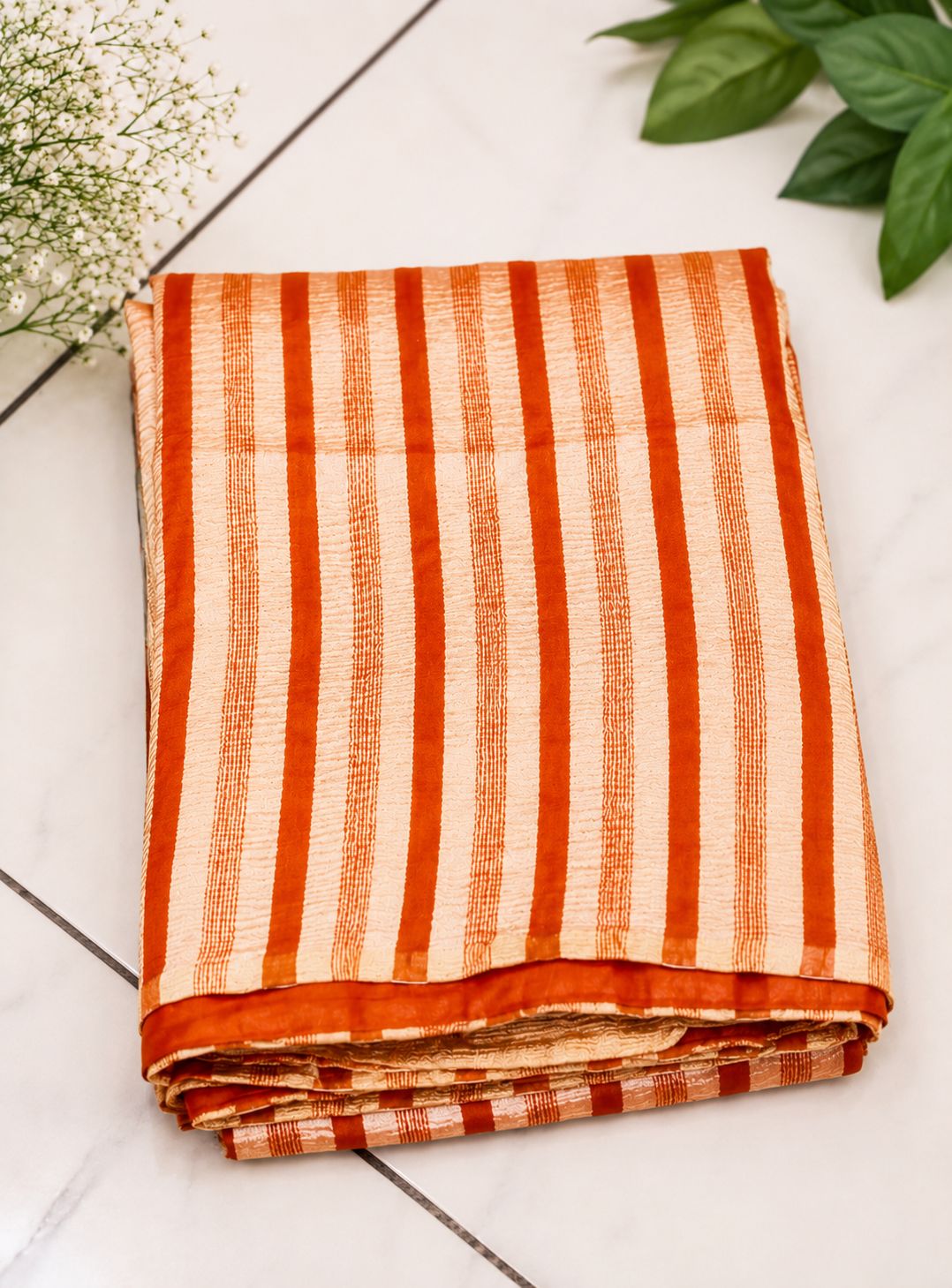 Folded fabric with orange and white stripes on a light wooden surface with green leaves.
